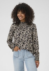 Blouse à manches longues avec un col froncé, présentant un motif zigzag noir et beige, fabriquée en tissu léger. Associée à un jean délavé.
