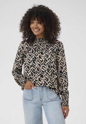 Blouse à manches longues avec un col froncé, présentant un motif zigzag noir et beige, fabriquée en tissu léger. Associée à un jean délavé.