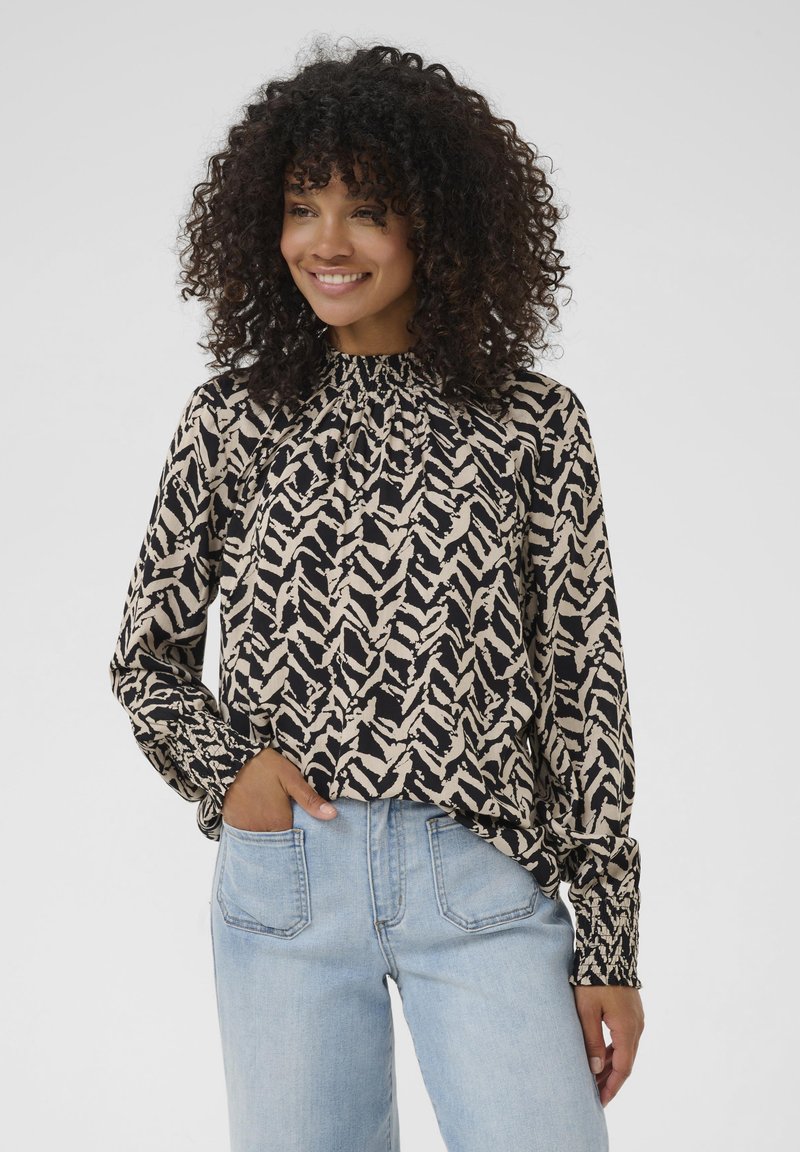 Blouse à manches longues avec un col froncé, présentant un motif zigzag noir et beige, fabriquée en tissu léger. Associée à un jean délavé.