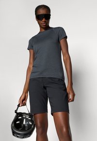 Herre atletisk antrekk med en tettsittende mørk grå skjorte og svarte shorts. Modellen holder en svart hjelm og har på seg store, mørke solbriller.