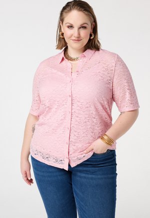 Vrouw die een roze kanten knoopblouse draagt, blauwe jeans, gouden oorringen, gelaagde kettingen en gouden armbanden, poserend met één hand in de zak.