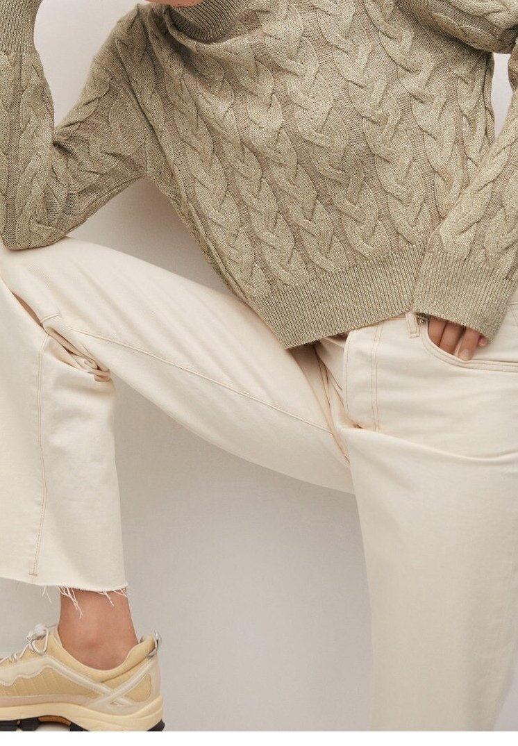 Personne portant un pull en tricot beige à torsades, un pantalon court blanc cassé et des baskets beiges, posant avec une jambe pliée et la main dans la poche.