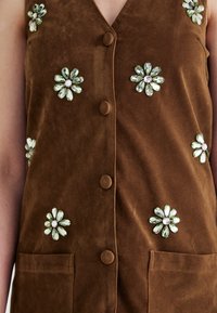 Brown suede vest with a V-neck, featuring five floral embellishments of green and clear gems, and two front pockets. Button closure detail.