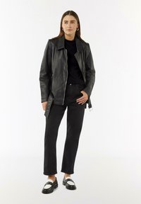 Zwarte leren jas met een kraag, ritssluiting en twee zijzakken, gecombineerd met zwarte broek en zwart-witte loafers.