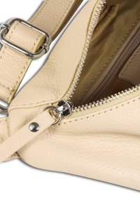 Beige leren tas met textuur, close-up, met een metalen ritssluiting, gestikte randen en een verstelbare band met een zilverkleurig metalen detail.