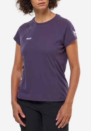 Frau trägt ein lila kurzärmeliges Sportshirt von Millet und schwarze Hose, steht mit entspannten Händen neben dem Körper.