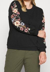 Czarny sweter z kolorową haftowaną florą na rękawach, z ściągaczami na mankietach i u dołu. W komplecie z oliwkowymi spodniami.
