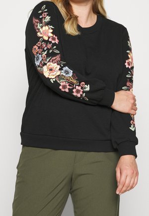 Czarny sweter z kolorową haftowaną florą na rękawach, z ściągaczami na mankietach i u dołu. W komplecie z oliwkowymi spodniami.