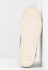 Semelle d'une basket blanche à rainures horizontales avec le logo "REPLAY", positionnée verticalement contre un mur d'angle blanc.