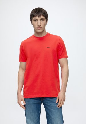 Jeune homme portant un T-shirt BOSS rouge vif et un jean bleu, debout devant un fond blanc uni, face à l'avant.