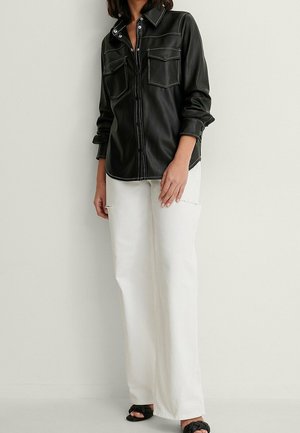 Femme portant une chemise noire en cuir à manches longues avec des poches avant, un pantalon blanc à jambes larges, et des talons ouverts tissés noirs, debout contre un mur blanc.