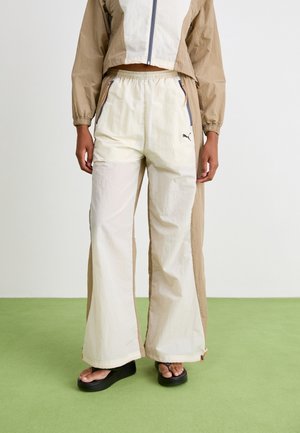 PUMA windbreaker en lichtgekleurde, wijde broek met ritszakken en contrasterende zijpanelen, gedragen door een model in platformsandalen.