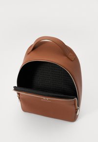 Mochila de imitación cuero marrón con forma redondeada, asa superior y cremallera plateada. El interior presenta un forro de tela negra con un patrón de logotipos.