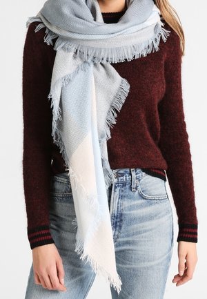 Femme portant un pull bordeaux, un jean bleu clair et une grande écharpe à franges aux tons pastel bleu et crème enroulée autour du cou.