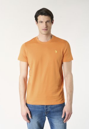 Uomo che indossa una maglietta arancione semplice a maniche corte con un piccolo logo giallo sul petto e jeans blu, in piedi davanti a uno sfondo bianco.