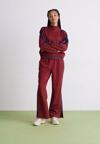 Bordeaux tracksuit med en lynlåslukket jakke med marineblå detaljer og matchende bukser med sideslidser. Båret med lyse sneakers.