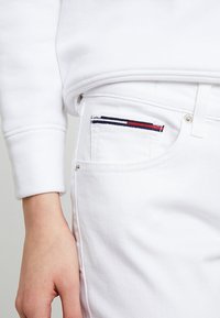 Jeans de mezclilla blanca con un bolsillo frontal que presenta un acento a rayas en marino, rojo y blanco. Textura suave y diseño ajustado.