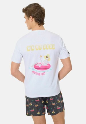 Hombre con camiseta blanca con un esqueleto con sombrero de sol flotando en un tubo rosa, sosteniendo una bebida, con el texto "ESTAMOS BIEN" y "EL POSITIVISMO PRIMERO."