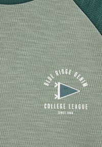 Zöld textúrájú szövet fehér felirattal, melyen az áll: "Blue Ridge Denim College League Since 1986", valamint egy kis háromszög alakú zászlólogó.