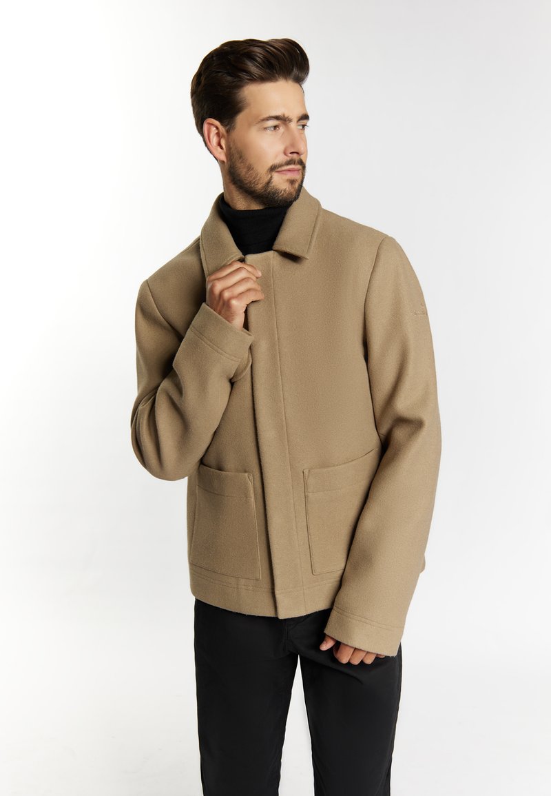 Jachetă din amestec de lână bej, cu guler, buzunare frontale și un design drept. Asortată cu o bluză de tip turtleneck neagră și pantaloni închiși la culoare.