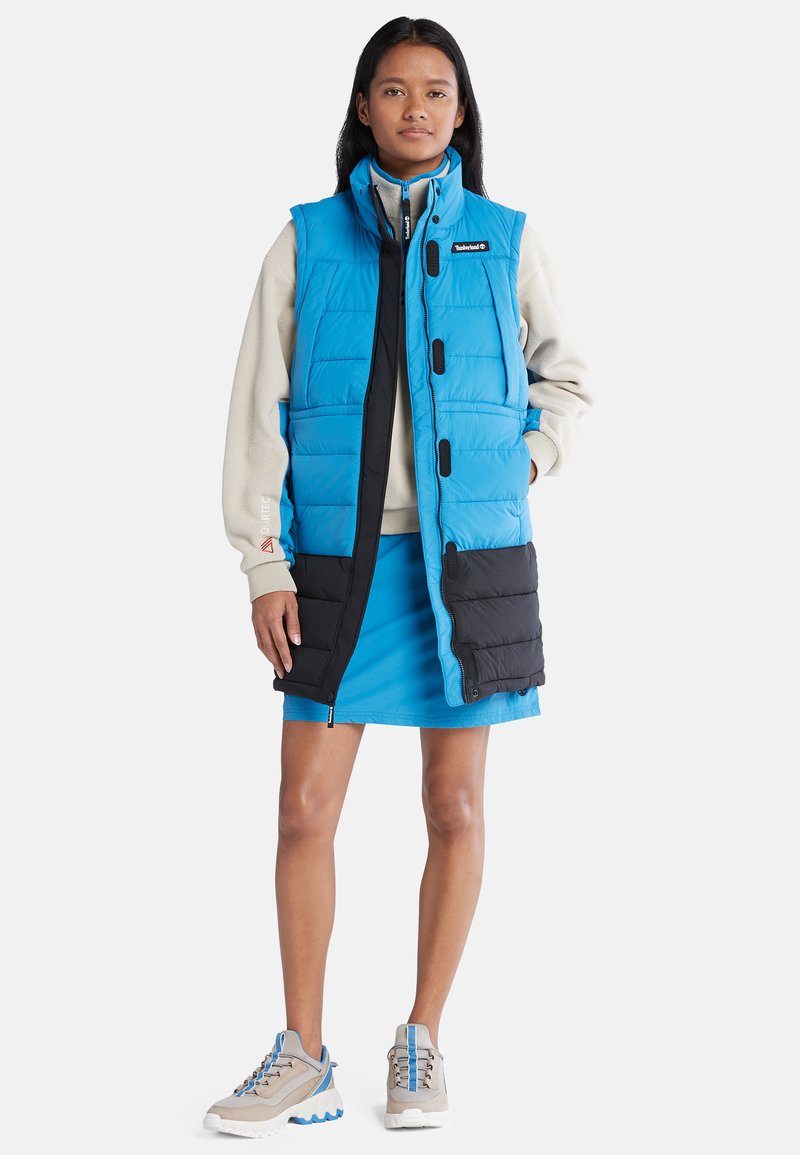 Blau- und schwarz gepolsterte Weste mit hohem Kragen, die über einem hellgrauen Fleecepullover und einem blauen Rock getragen wird. Beigefarbene Sneakers mit blauen Akzenten.