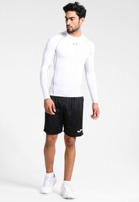 Mężczyzna stoi, ubrany w przylegający biały sportowy top z długim rękawem, czarne sportowe spodenki i białe tenisówki na jednolitym białym tle.