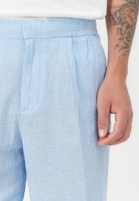 Light blue trousers made of textured fabric with vertical white stripes, featuring a flat front, side pockets, and an elasticated waist.
