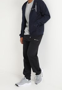 Felpa con cappuccio blu navy con zip, t-shirt grigia, pantaloni sportivi neri e scarpe sportive grigie con accenti neri. La felpa ha coulisse e logo.