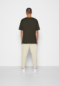 Camiseta de manga corta verde oscuro con un corte relajado, combinada con pantalones beige claros ajustados y zapatillas blancas, vista desde atrás.
