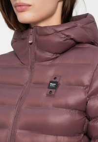 Zateplená bunda v tlmenej mauve farbe, s vysokým golierom, zipsom a logom na hrudi, doplnená o zapínanie na cvoky.
