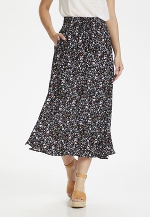 Femme portant une jupe midi noire à motif floral avec poches, associée à des sandales compensées marron à bride cheville et un haut blanc.