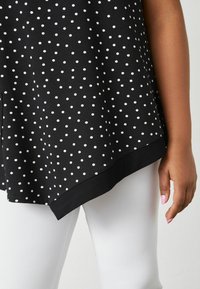 Top nero con pois bianchi, orlo curvo, realizzato in tessuto morbido. Abbinato a leggings bianchi a coste, che mostrano un primo piano del tessuto.