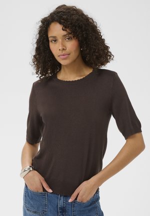 Femme aux cheveux bouclés portant un pull à manches courtes marron foncé et un jean bleu, debout les mains dans les poches avant, sur fond blanc.