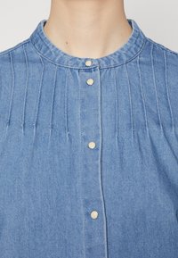 Šviesiai mėlyna džinsinė marškinė su stovinčiu apykakle, vertikaliomis raukšlėmis ir perlamutrinėmis spaudėmis priekyje. Minkšta tekstūra.