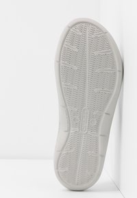 Semelle en caoutchouc blanche avec un motif en zigzag texturé et des points surélevés, intégrant un détail de logo. Le profil latéral montre le contour et le design léger.