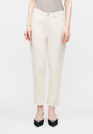 Weibliches Model trägt hochtaillierte, gerade geschnittene Jeans in Off-White, kombiniert mit schwarzen spitz zulaufenden High Heels und einem grauen ärmellosen Oberteil.