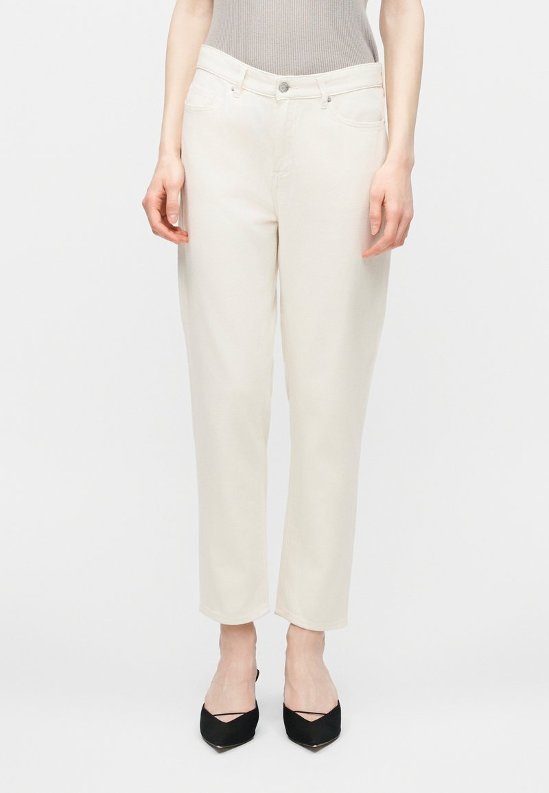 Γυναίκα μοντέλο που φορά ψηλόμεσα, εκρού τζιν με ίσια μπατζάκια, συνδυασμένα με μαύρες μυτερές γόβες και γκρι αμάνικο τοπ.