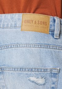 Svetlo modri kavbojke z raztrganinami na zadnjem žepu. Oblečen usnjen etiket s napisom "ONLY & SONS" in "PASSIONATE JEANS MAKERS."