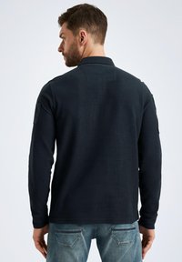 Marineblaues Langarm-Polohemd aus strukturiertem Stoff mit Kragen und Schulterpatch-Details, kombiniert mit blauen Jeans. Rückansicht gezeigt.