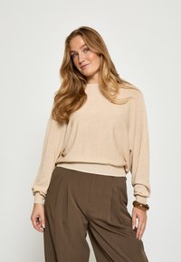 Gai bēša džemperis ar garām, brīvām piedurknēm un ribotu apakšmalu, apvienots ar brūnām, pleitētām biksēm ar augstu jostasvietu. Gluda, mīksta tekstūra.