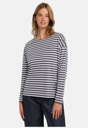 Frau trägt ein grau-weiß gestreiftes Langarmshirt mit horizontalen Streifen und dunkle Jeans, steht vor einem schlichten hellen Hintergrund.