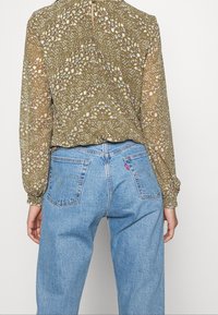 Olivgrüne Bluse mit floralen Muster, Knopfverschluss am Hals, lange Ärmel, kombiniert mit hellblauen, hochgeschnittenen Jeans.