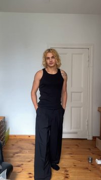 Top preto sem mangas e calças largas pretas. Tecido suave com um ajuste descontraído. Cabelo loiro levemente texturizado, em pé no interior com um piso de madeira.