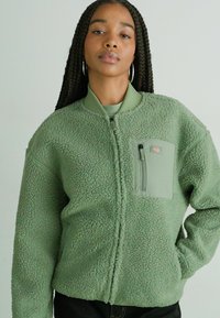 Green fleece bomber jacket with a textured surface, full zip, ribbed collar, and a zippered chest pocket in a smooth fabric.