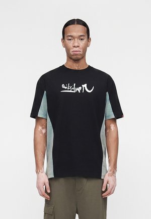 Jeune homme atteint de vitiligo porte un t-shirt Quiksilver noir et gris et un pantalon cargo olive, debout devant un fond blanc uni.