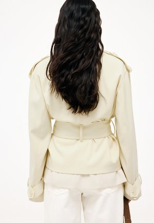 Mujer con cabello largo y oscuro ondulado, vistiendo una chaqueta beige con cinturón y pantalones blancos, de pie de espaldas a la cámara contra un fondo liso.