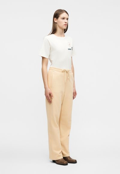 Νέα γυναίκα στέκεται φορώντας λευκό κοντομάνικο μπλουζάκι με σχέδια, μπεζ παντελόνια με κορδόνι και καφέ παπούτσια με τακούνι σε απλό φόντο.