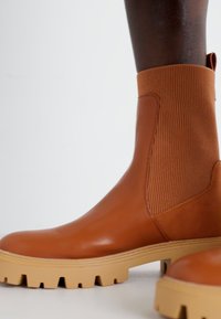 Botte cheville en cuir marron avec panneau textile côtelé, semelle en caoutchouc épaisse crème et finition lisse. Anse latérale pour un enfilage facile.