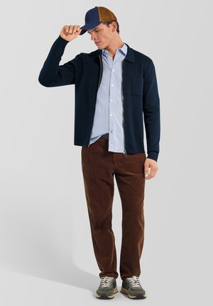 Hombre con gorra azul y marrón, chaqueta azul marino con cremallera, camisa azul clara abotonada, pantalones de pana marrones y zapatillas grises, de pie sobre un fondo gris.