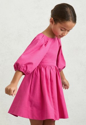 Noor tüdruk tume juustega madalas kuklis, kandes erkroosa kleiti põrutavatele varrukatele, vaadates alla ja kergelt käsi sirutades.